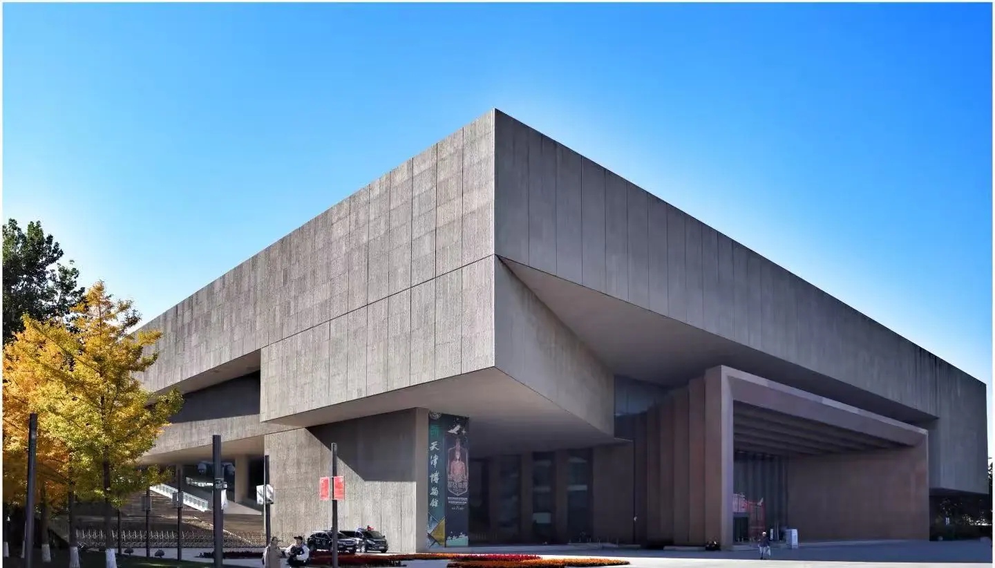 Large modern concrete building with a cantilevered upper section, glass entrance, and autumn trees on the left under a clear blue sky.
