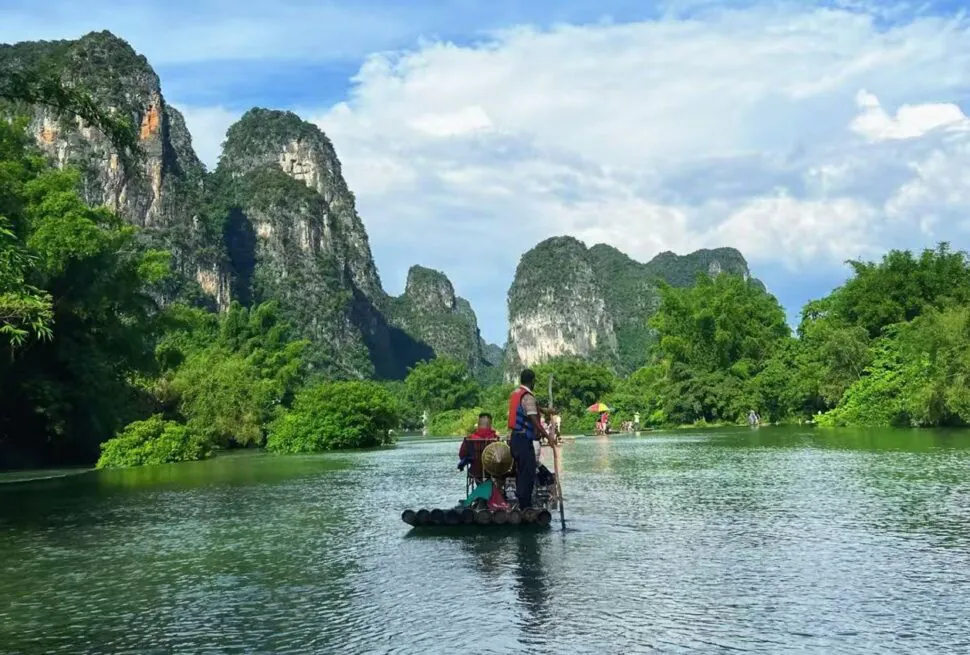 People paddling on a bamboo raft along a calm river, surrounded by lush green trees and towering rocky cliffs under a blue sky.Bamboo Rafting on the Yulong River,Yangshuo,Guangxi,Chinatravel