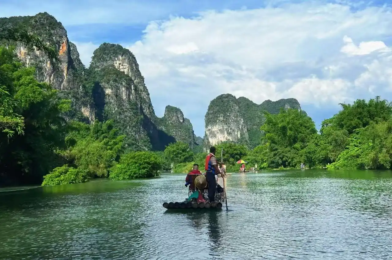 People paddling on a bamboo raft along a calm river, surrounded by lush green trees and towering rocky cliffs under a blue sky.Bamboo Rafting on the Yulong River,Yangshuo,Guangxi,Chinatravel
