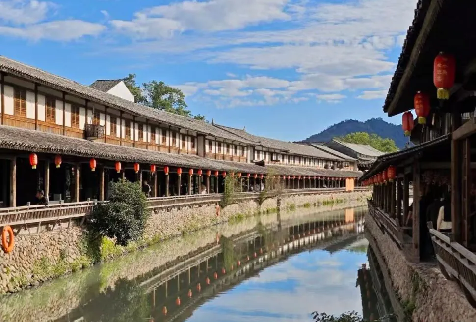 Yantou Ancient Village ,Wenzhou,Chinatour Row of traditional wooden shops along a calm canal, red lanterns hanging overhead, blue sky and hills in the background.