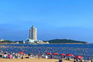 Best Beaches,Qingdao,Chinatour Crowded beach with people in shallow water, red umbrellas on the sand, and a tall pyramid-topped building in the distance against a blue sky.