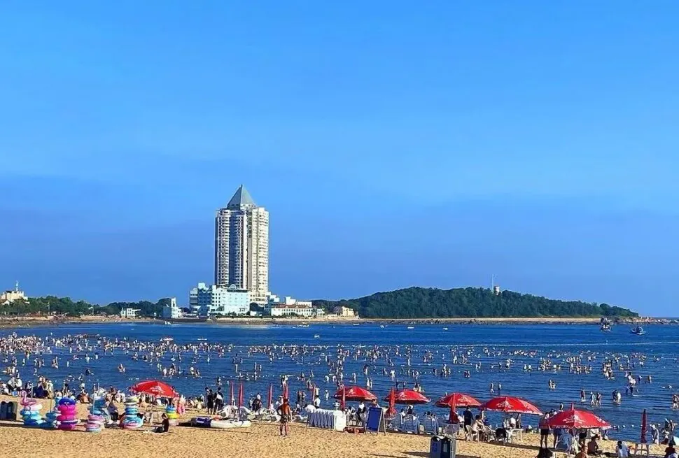 Crowded beach with people in shallow water, red umbrellas on the sand, and a tall pyramid-topped building in the distance against a blue sky.