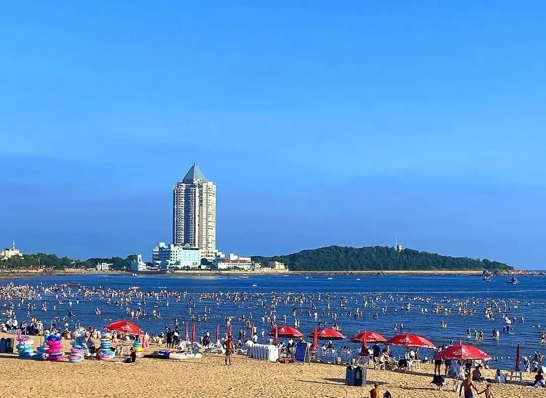 Best Beaches,Qingdao,Chinatour Crowded beach with people in shallow water, red umbrellas on the sand, and a tall pyramid-topped building in the distance against a blue sky.