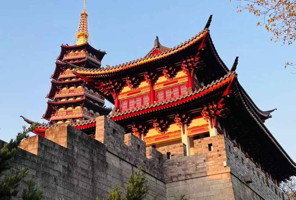 Wuzhou Ancient City,Jinhua,Chinatour Red and black multi-tiered Chinese pagoda atop a stone fortress wall against a clear blue sky, with ornate curved roofs and golden pinnacle.
