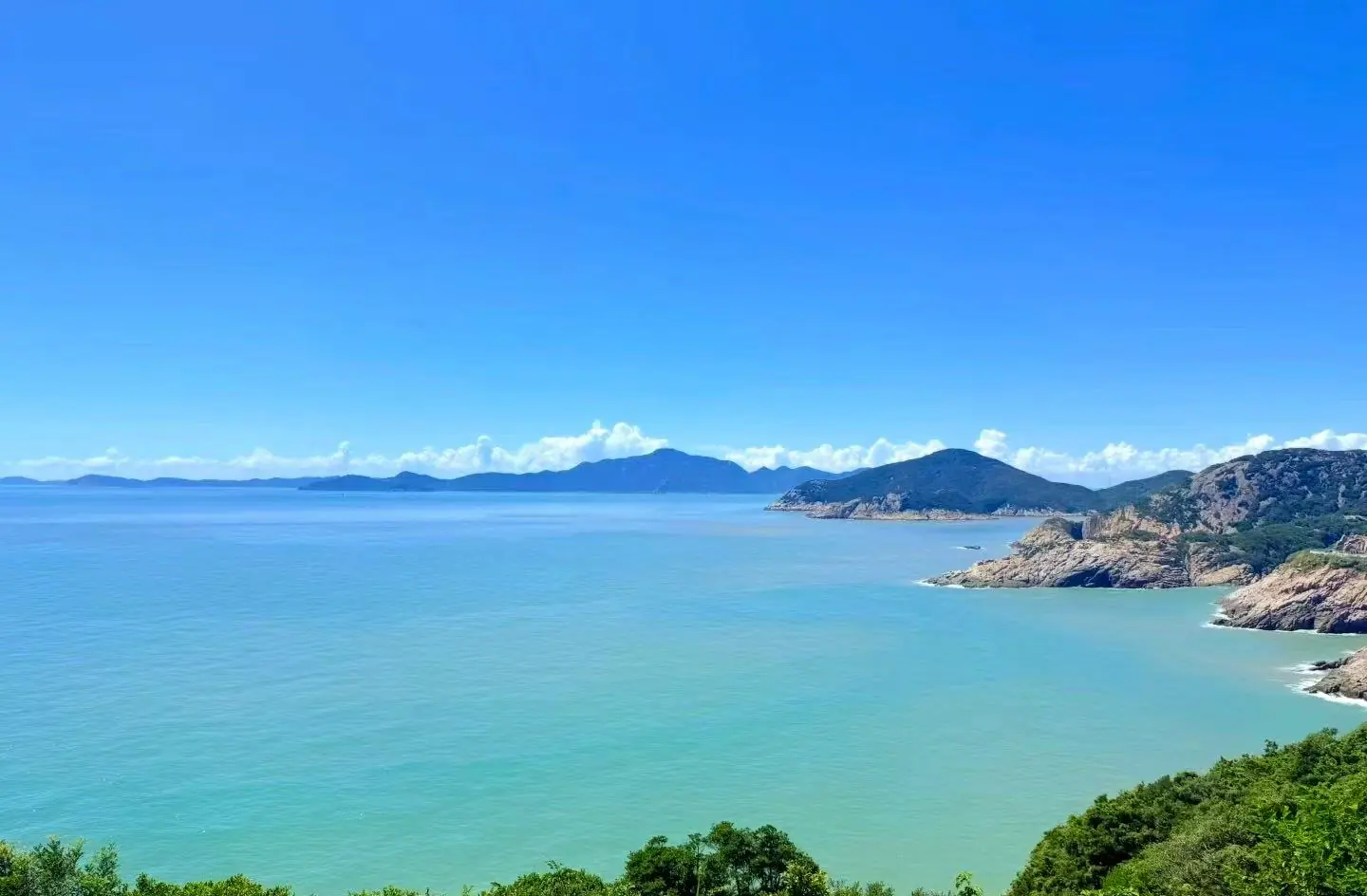 Zhujiajian Island,Zhoushan,Chinatour, Bright coastal landscape with turquoise water, rocky cliffs, and green hills under a clear blue sky, conveying serenity and openness.