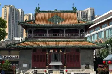Traditional Chinese temple with red doors and ornate green-tiled roof, set against modern high-rises in an urban plaza.