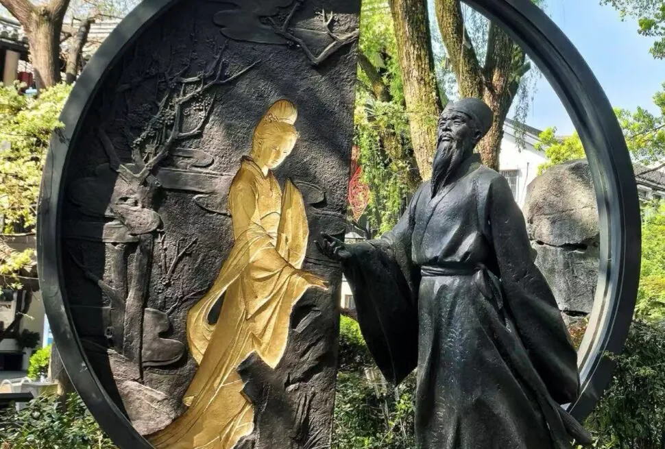 Shen Garden ,Shaoxing,Chinatour Circular sculpture featuring a gold-toned seated female figure and a dark-robed man in a garden setting, with a large circular frame surrounding them.
