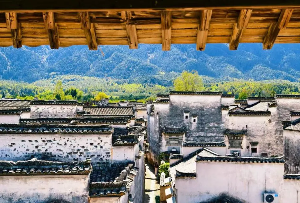 Nanping Ancient Town,Huangshan,Chinatour Traditional Chinese village with white-walled houses and tiled roofs framed by a wooden balcony, against green hills and mountains.