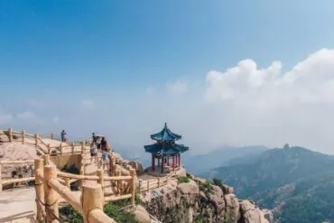 Mount Laoshan Scenic Area ,Qingdao,Chinatour Hikers walk along a wooden railing on a rocky mountain path, with a small blue-roofed pavilion perched on the rocks.