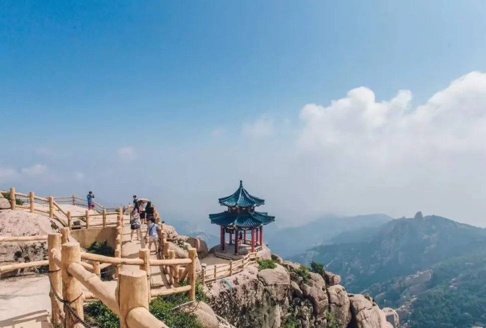 Mount Laoshan Scenic Area ,Qingdao,Chinatour Hikers walk along a wooden railing on a rocky mountain path, with a small blue-roofed pavilion perched on the rocks.