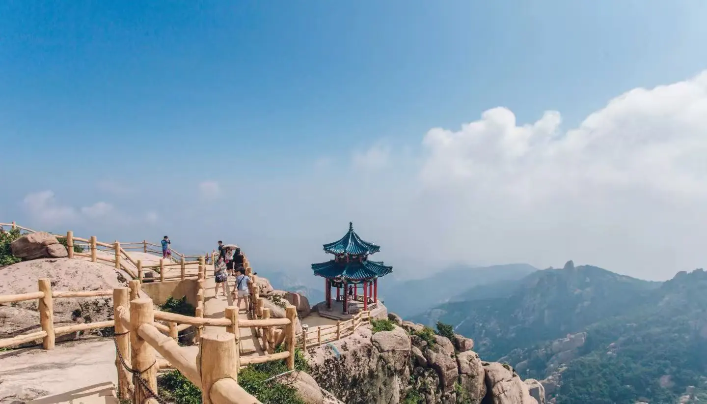 Mount Laoshan Scenic Area ,Qingdao,Chinatour Hikers walk along a wooden railing on a rocky mountain path, with a small blue-roofed pavilion perched on the rocks.