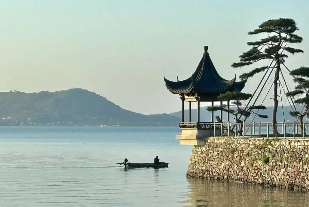 Dongqian Lake,Ningbo,Chinatour Lakeside scene with a pagoda-style pavilion on a stone platform and two people rowing a small boat on calm water, hills in the distance.