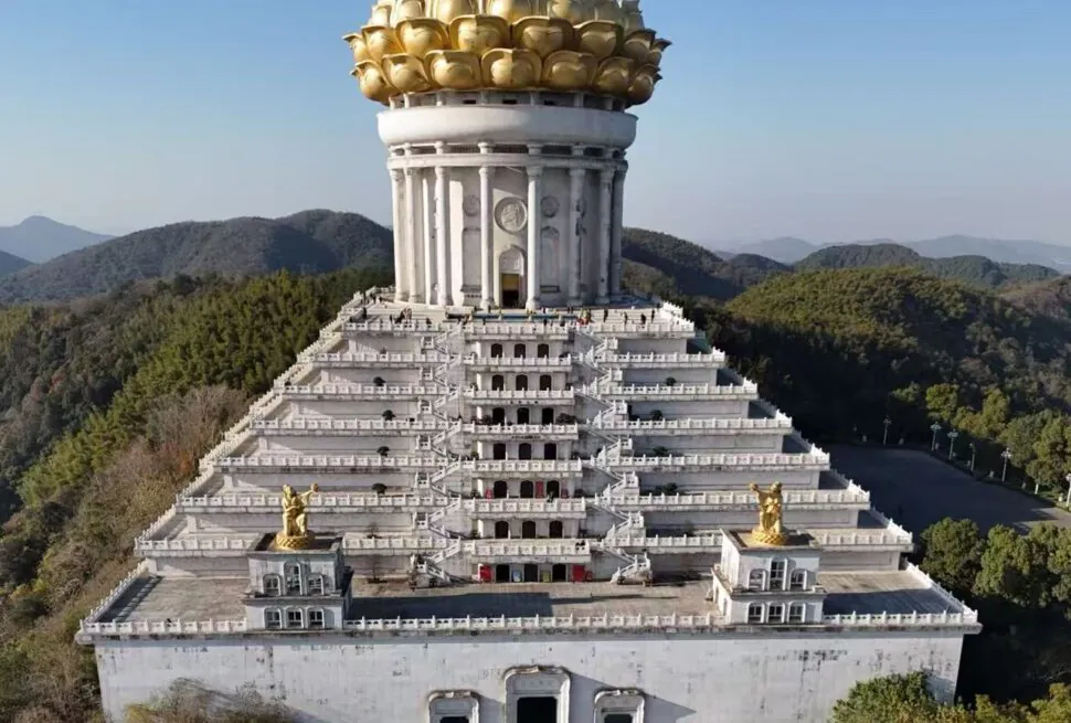A giant white tiered temple on a hill, topped with a gilded lotus crown and surrounded by forested mountains.