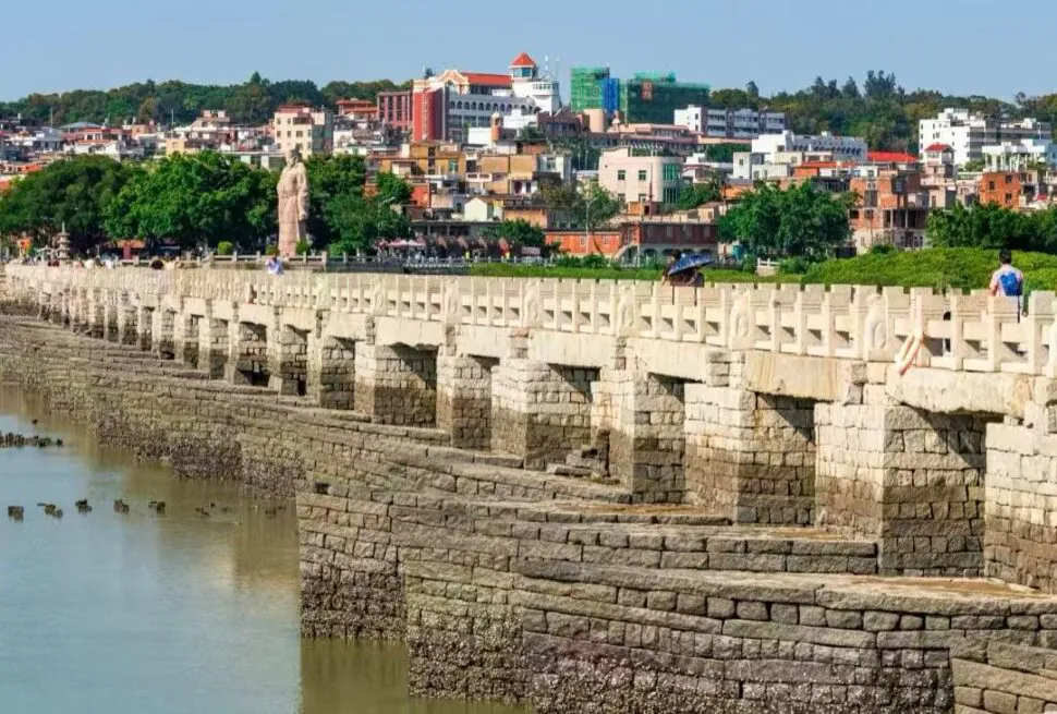 Stone arched bridge along a calm river, with a town and green trees in the background; people stroll the pedestrian walkway under a clear sky.