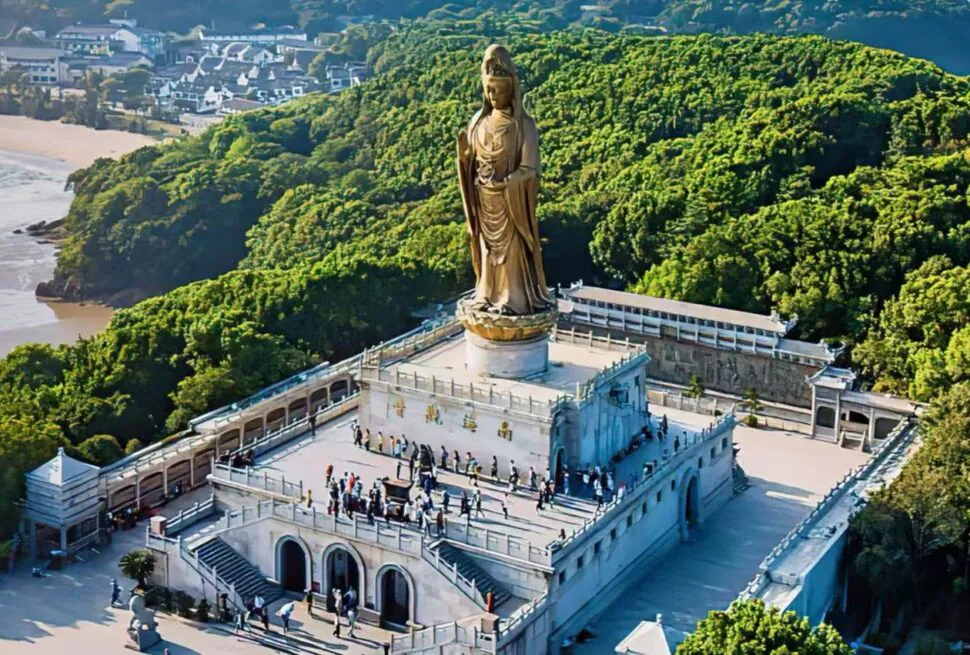 Putuo Mountain,Zhoushan,Chinatour A tall golden statue on a pedestal atop a white temple complex, surrounded by greenery with visitors on the terraces below.