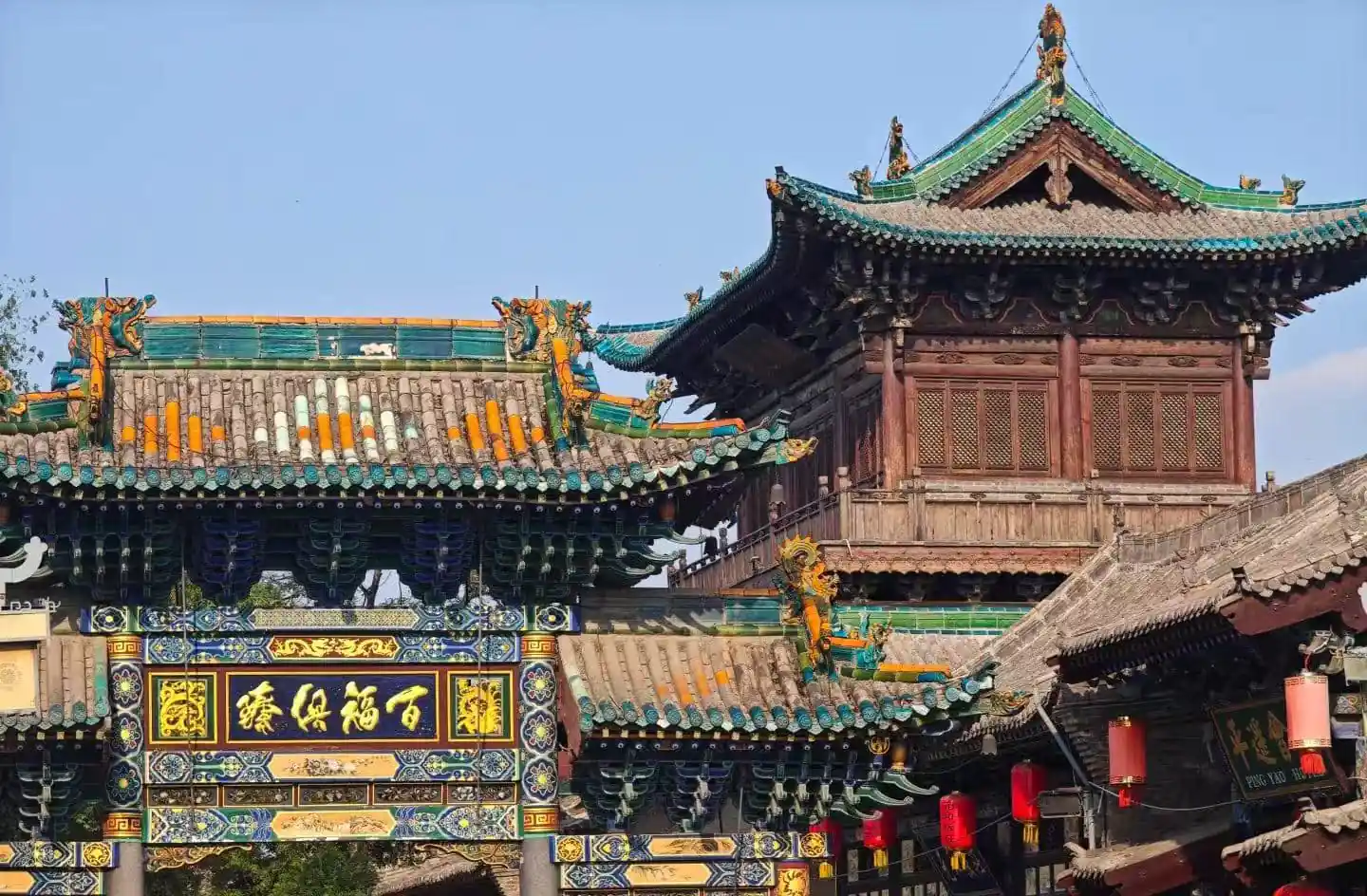 Pingyao City God Temple,Pingyao,Chinatour Ornate Chinese temple gate with colorful tiled roof, carved wood, and a decorative plaque in Chinese characters under a bright blue sky.