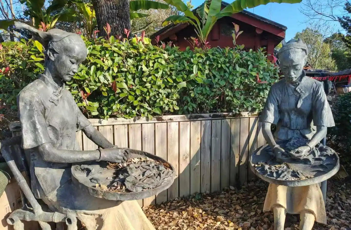 Wuyi Tea Expo Park,Nanping,Chinatour Two life-size stone statues of women in traditional outfits, each holding a round tray filled with rocks, set in a garden with a wooden fence and leafy plants behind them