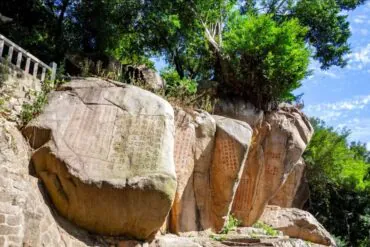 Jiuri Mountain Wind-praying Stone Inscriptions,Quanzhou,Chinatour Large boulder with Chinese inscriptions carved on its face, set among rocks and trees on a sunny hillside.