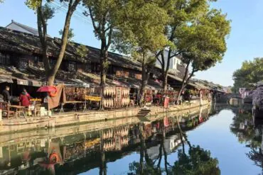 Anchang Ancient Town ,Shaoxing,Chinatour Canal-side street with traditional buildings, market stalls, and trees, reflected in calm water beneath a clear blue sky.