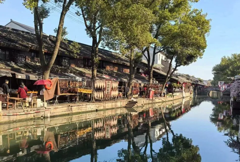 Canal-side street with traditional buildings, market stalls, and trees, reflected in calm water beneath a clear blue sky.