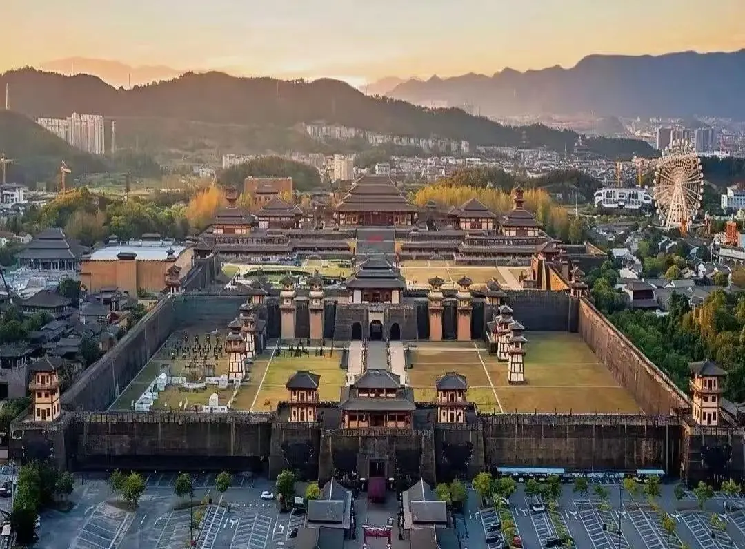 Hengdian World Studios ,Jinhua,Chinatour Sprawling ancient Chinese fortress with tall stone walls, pagoda roofs, and inner courtyards at sunset; city and hills in the background.