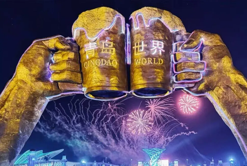 Night view of giant golden fists forming an arch with the words 'QINGDAO WORLD' and colorful fireworks above a lit cityscape outside the venue.