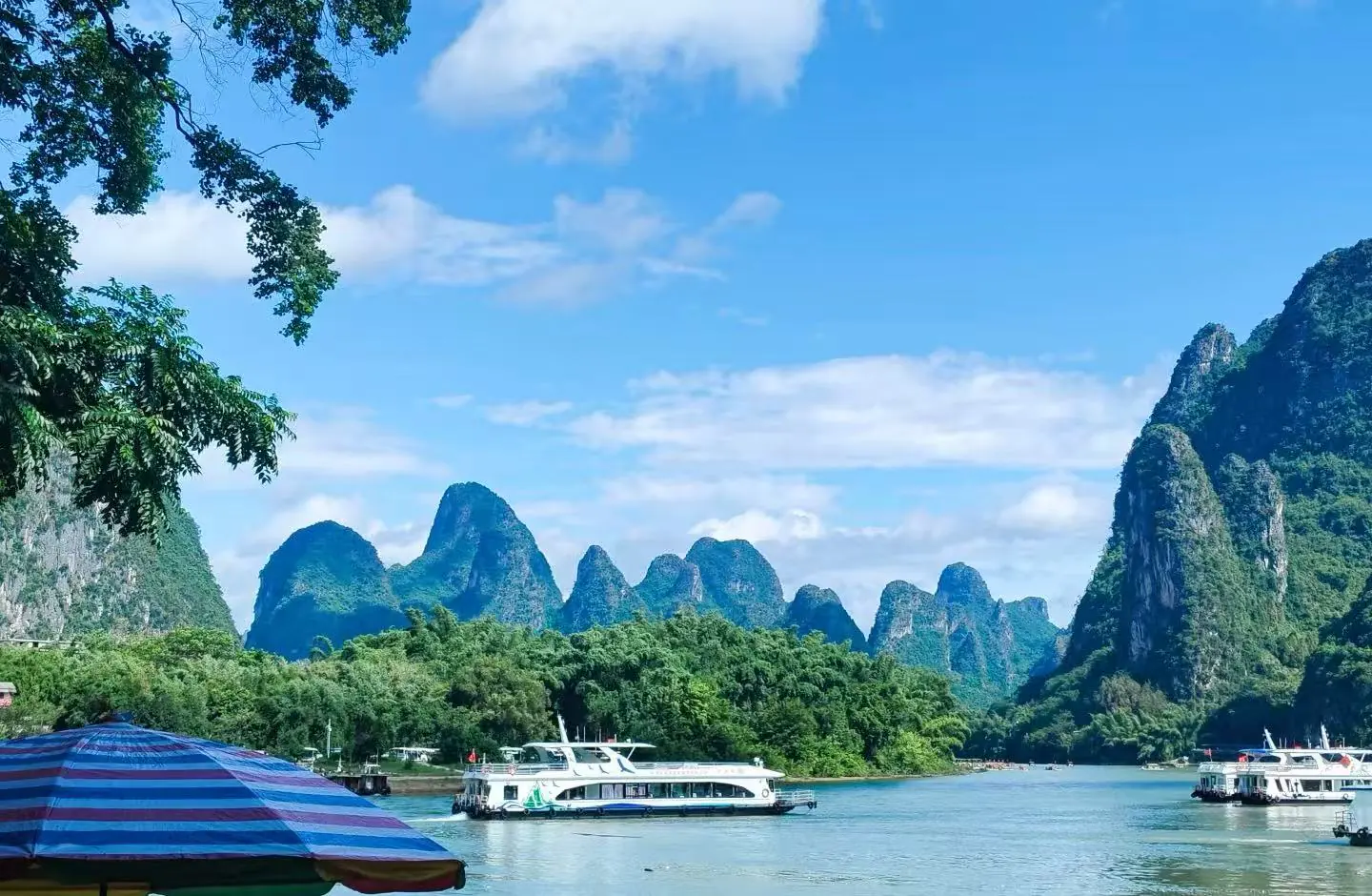 Scenic river lined with lush green trees and towering limestone karst hills under a blue sky; boats docked along the waterfront and a striped umbrella in the foreground.Xingping Ancient Town,Yangshuo,Guangxi,Chinatravel