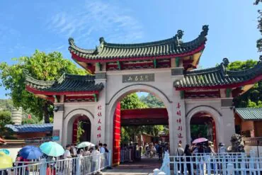 Nanputuo Temple,Xiamen,Chinatour Traditional Chinese temple entrance with curved roofs and red columns; people with umbrellas queue to enter on a sunny day.