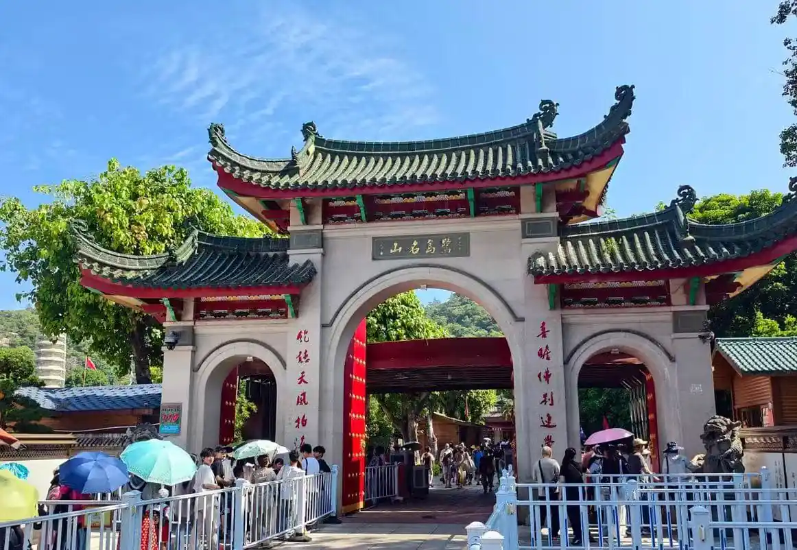 Nanputuo Temple,Xiamen,Chinatour Traditional Chinese temple entrance with curved roofs and red columns; people with umbrellas queue to enter on a sunny day.
