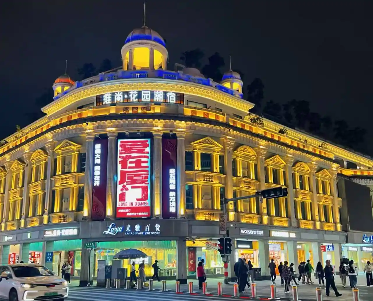 Zhongshan Road Pedestrian Street,Xiamen,Chinatour Nighttime view of a grand, neoclassical-style building illuminated in warm yellow lights, with a large red vertical billboard and people crossing the street at a busy intersection.