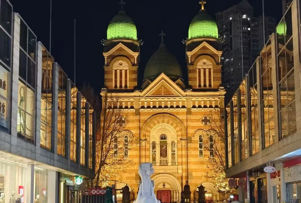St. Joseph Cathedral Xikai Church,Tianjin,Chinatour Night view of a brick church with two green-domed towers at a busy city street, framed by glass storefronts and warm lights.