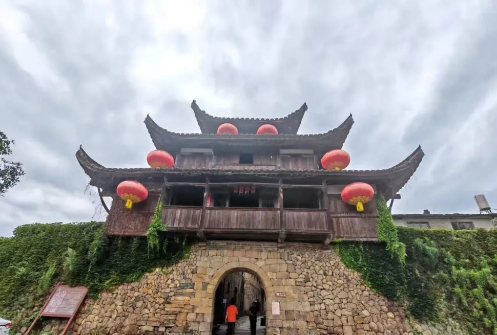 Traditional wooden gate tower atop a stone archway, adorned with red lanterns and creeping ivy against a cloudy sky treated as an historic entrance.Hepingi Ancient Town,Nanping,Fujian,Chinatravel