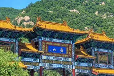 Ornate traditional Chinese gate with blue and gold decorative panels and orange tiled roofs, set against a green hillside mountain backdrop.