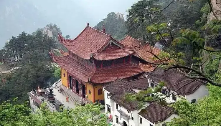 Temple complex with red-tiled roofs perched on a hillside, surrounded by trees and misty mountains.