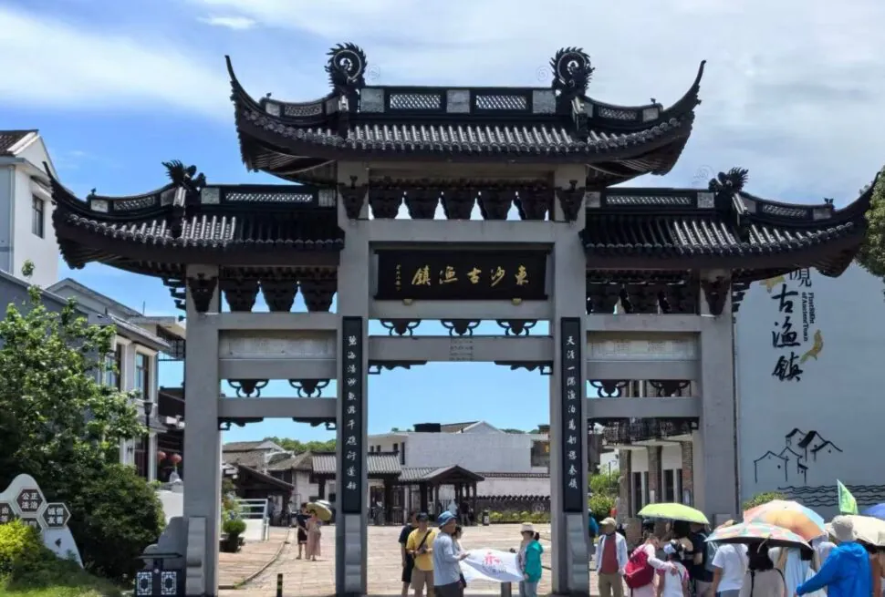 Dongsha Ancient Town,Zhoushan,Chinatour Traditional Chinese arch gate with ornate curved roofs and a dark plaque, visitors entering a cultural town under a blue sky.