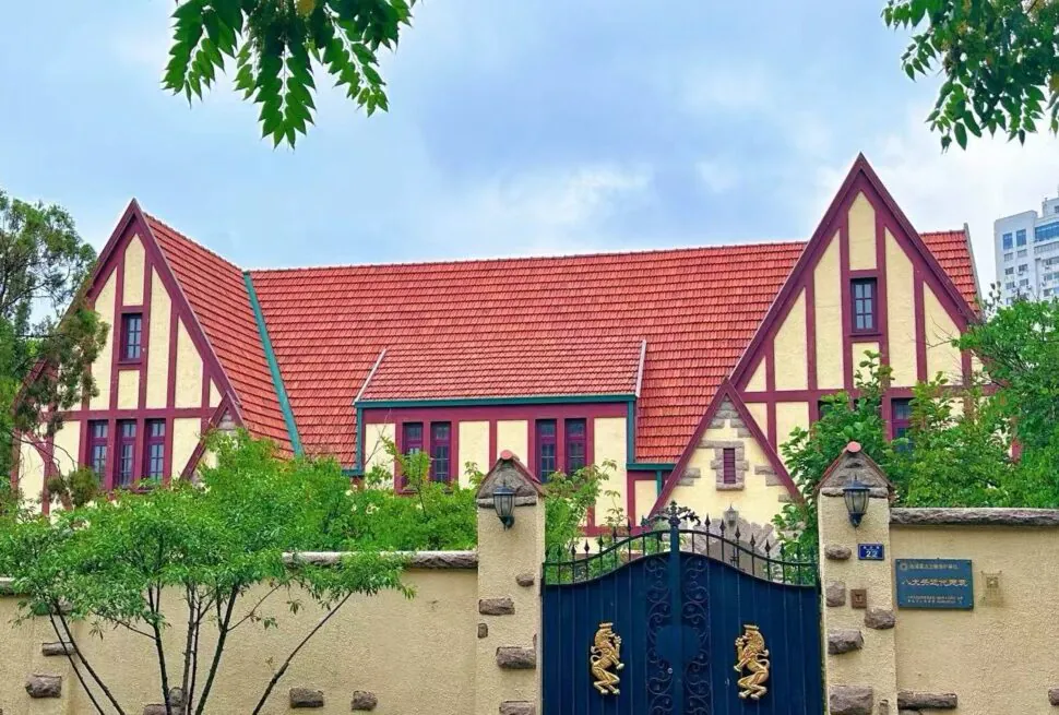 Colorful European-style house with yellow walls, red trim and steep red-tiled roof behind a stone fence and blue gate with dragon symbols, greenery in front.
