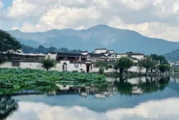 Tranquil riverside village with white-walled houses, a lotus-covered pond, and misty mountains in the background.
