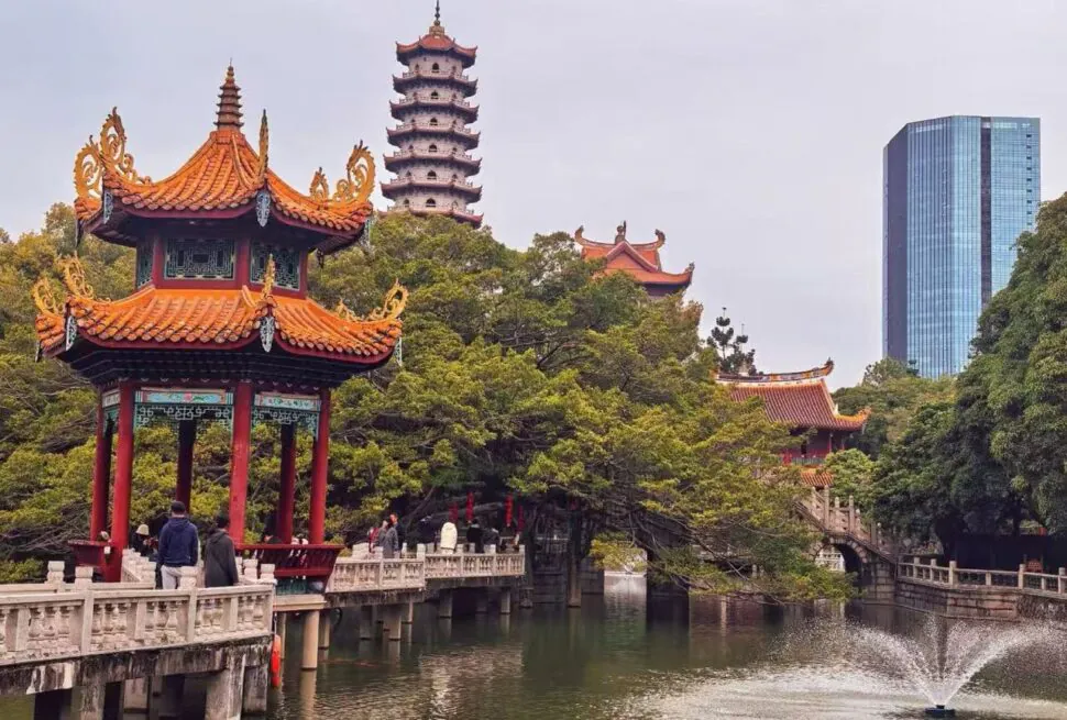 Red pagoda pavilion over a lake with a stone walkway and trees, a tall pagoda in the background and a modern glass building behind them.