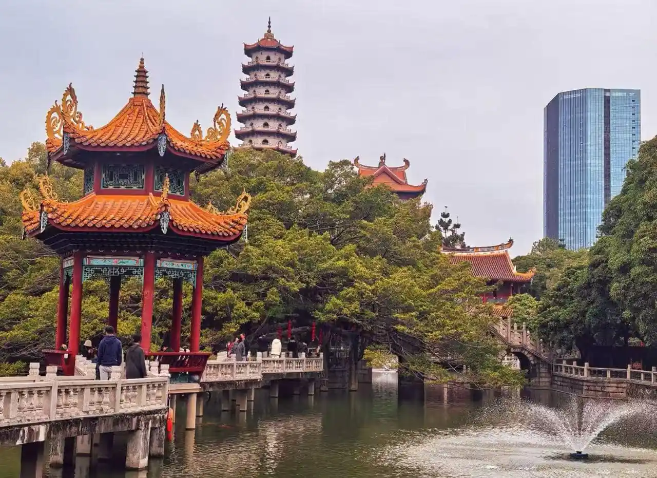 Xichan Temple,Fuzhou,Chinatour Red pagoda pavilion over a lake with a stone walkway and trees, a tall pagoda in the background and a modern glass building behind them.