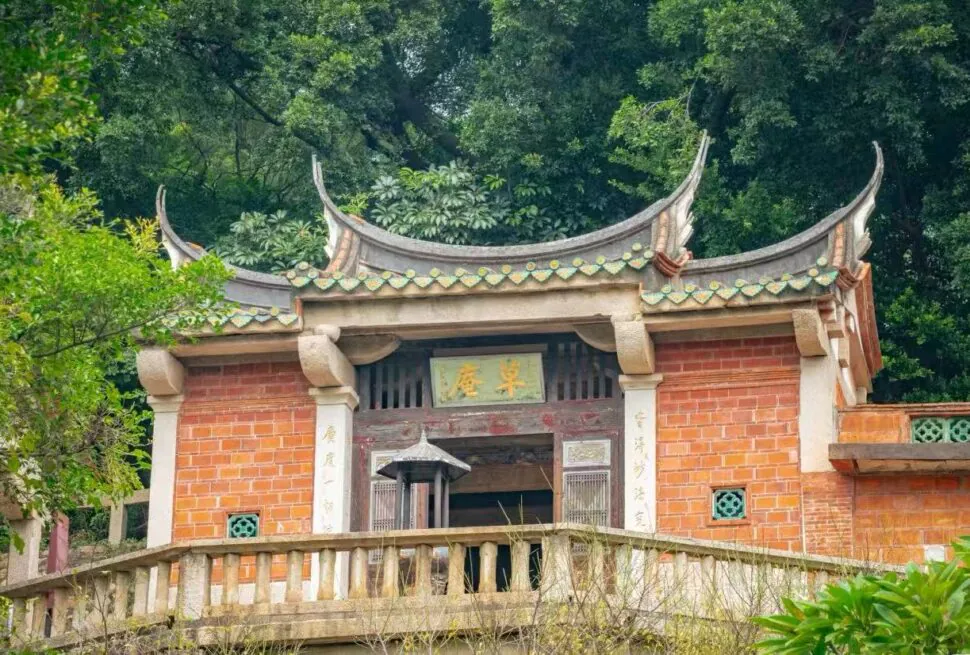 Grass Hut Manichaeism Site ,Quanzhou,Chinatour Traditional Chinese temple gate with curved tiled roof, red brick walls, and stone balustrade amid lush greenery