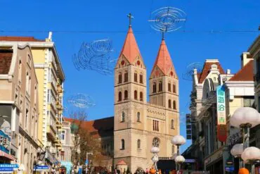 St. Michael’s Cathedral ,Qingdao,Chinatour Twin red-topped church towers rise over a busy city street under a clear blue sky, with spiral wire decorations overhead and storefronts on both sides.