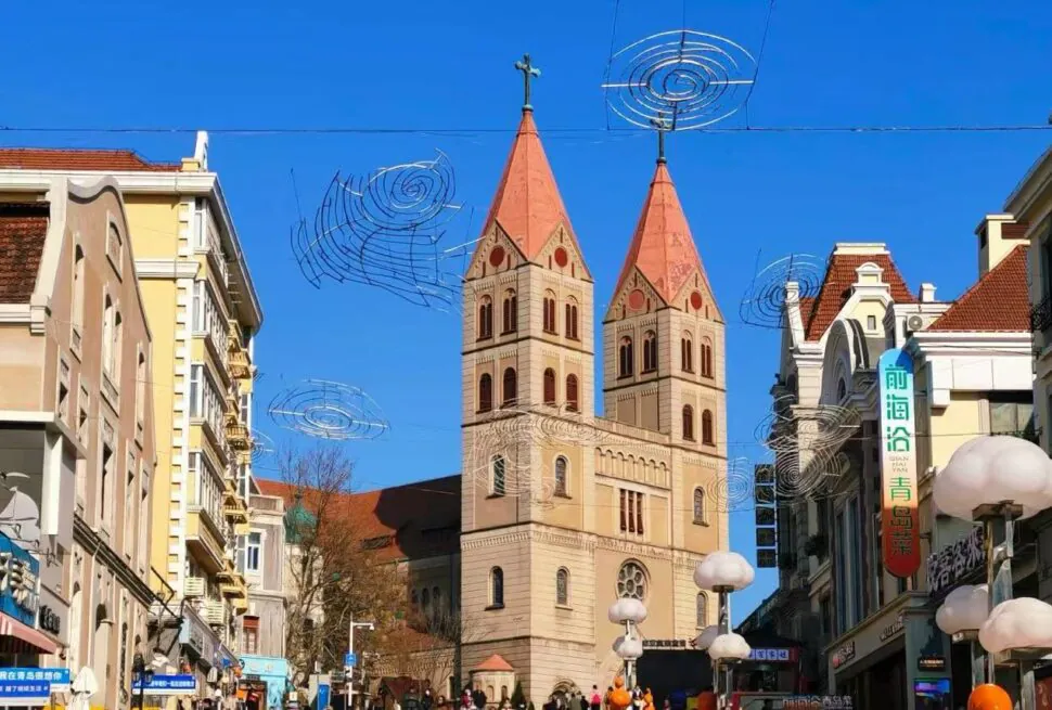 St. Michael’s Cathedral ,Qingdao,Chinatour Twin red-topped church towers rise over a busy city street under a clear blue sky, with spiral wire decorations overhead and storefronts on both sides.