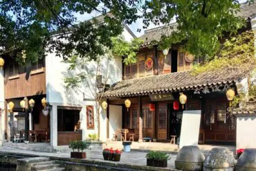 Yellow Wine Town ,Shaoxing,Chinatour Traditional Chinese riverside shopfronts with wooden lattice doors, yellow lanterns, and a tiled roof, along a narrow canal under leafy trees.