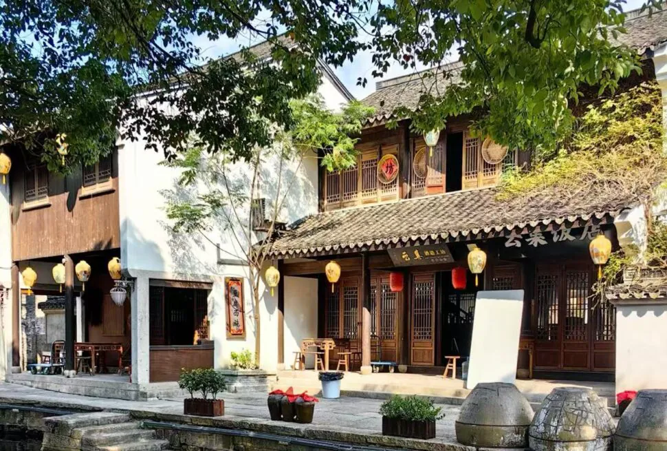 Traditional Chinese riverside shopfronts with wooden lattice doors, yellow lanterns, and a tiled roof, along a narrow canal under leafy trees.
