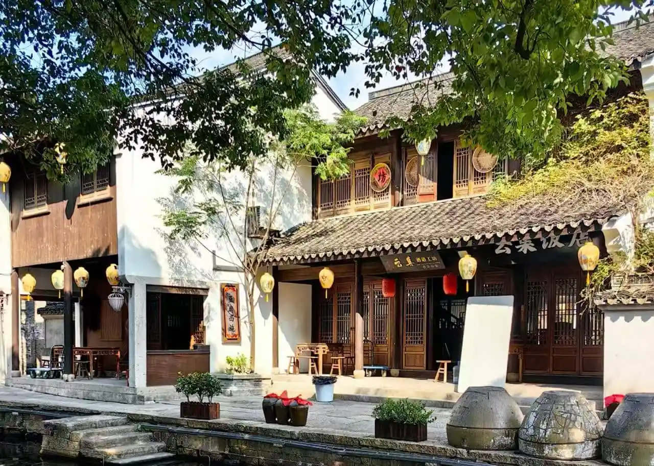 Yellow Wine Town ,Shaoxing,Chinatour Traditional Chinese riverside shopfronts with wooden lattice doors, yellow lanterns, and a tiled roof, along a narrow canal under leafy trees.