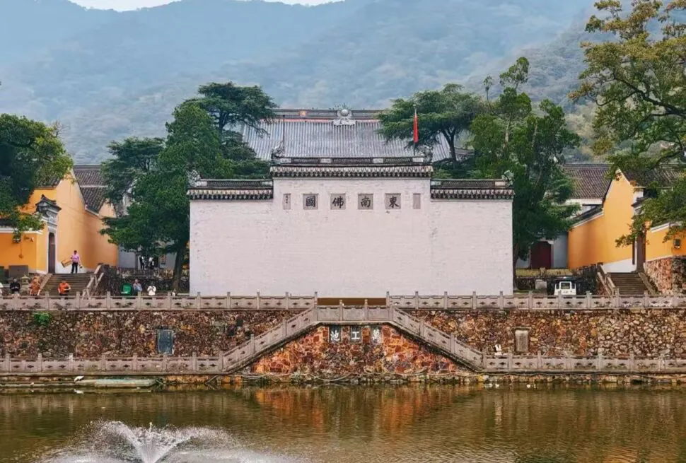 Temple complex by a pond with a white central wall, orange side buildings, and green trees against a mountainous backdrop.