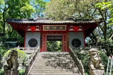 Great Buddha Temple ,Shaoxing,Chinatour Entrance to a traditional Chinese temple, red wooden gates atop stone steps, flanked by two stone guardian lion statues and hanging red lanterns in a lush garden setting.