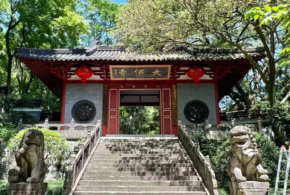 Great Buddha Temple ,Shaoxing,Chinatour Entrance to a traditional Chinese temple, red wooden gates atop stone steps, flanked by two stone guardian lion statues and hanging red lanterns in a lush garden setting.