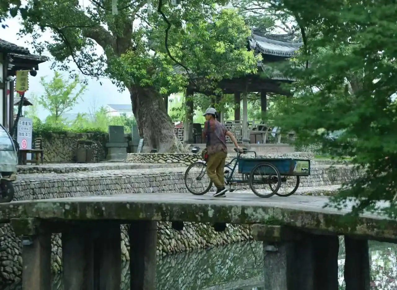 Nanxi River Scenic Area ,Wenzhou,Chinatour Man pushes a blue cargo tricycle across a stone-bridge over a canal, with trees and a pavilion in the background.