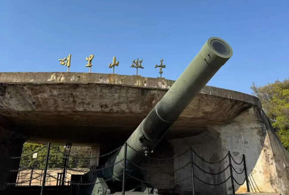 Military cannon protruding from a concrete bunker, angled upward, with blue sky and chain barriers around the display.